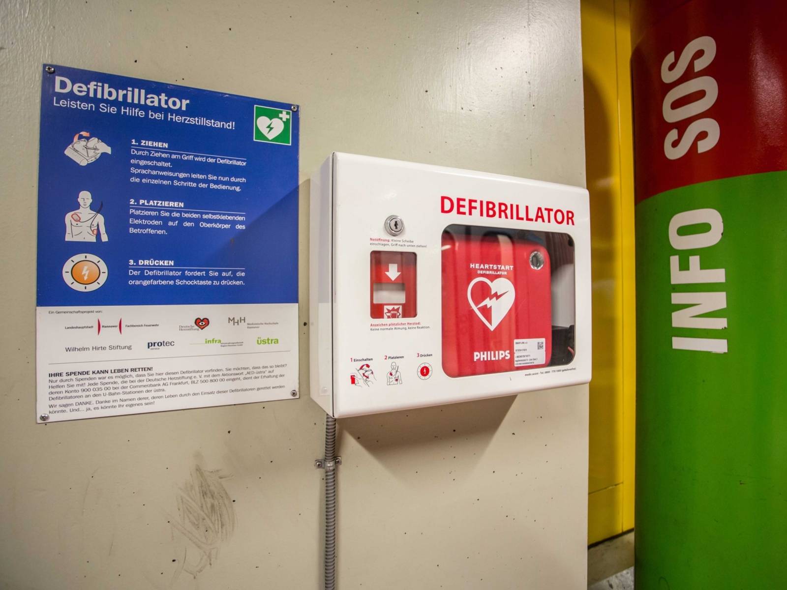 Ein Defibrillator an der Wand, daneben eine Notrufs&auml;ule