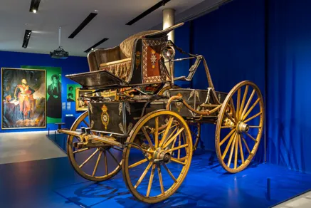 Blick in die Ausstellung "Vier Kutschen, ein Königreich. Hannover 1814 - 1866" , im Vordergrund ein Phaeton,  an der Wand Gemälde von Ernst August, König von Hannover und Ehefrau.