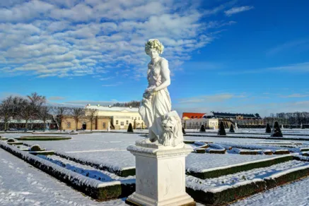 Schlossgarten im Winter mit einer schneebedeckten weißen Statue im Vordergrund und eleganten barocken Gebäuden im Hintergrund.