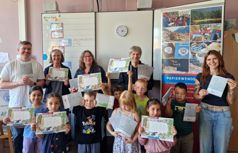 In einem Raum halten Menschen Stundenpläne und Schulhefte in den Händen und zeigen sie in die Kamera. In hinterer Reihe stehen fünf Erwachsene, vorne acht Kinder.