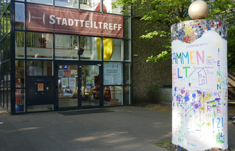 Vor dem Eingang des Stadtteiltreffs steht eine hohe bunt angemalte Litfaßsäule. Alle sind im Stadtteiltreff Willkommen!