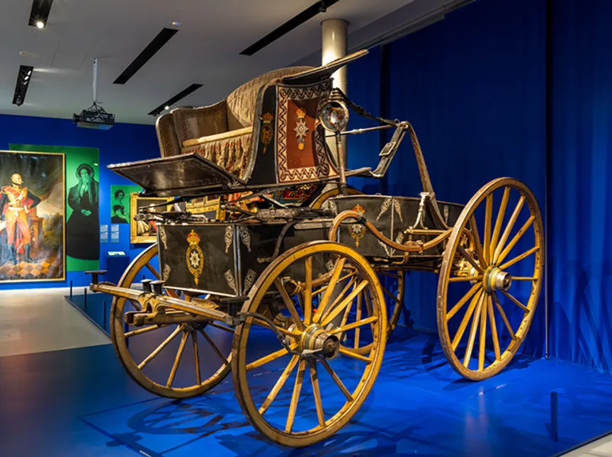 Blick in die Ausstellung "Vier Kutschen, ein Königreich. Hannover 1814 - 1866" , im Vordergrund ein Phaeton,  an der Wand Gemälde von Ernst August, König von Hannover und Ehefrau.